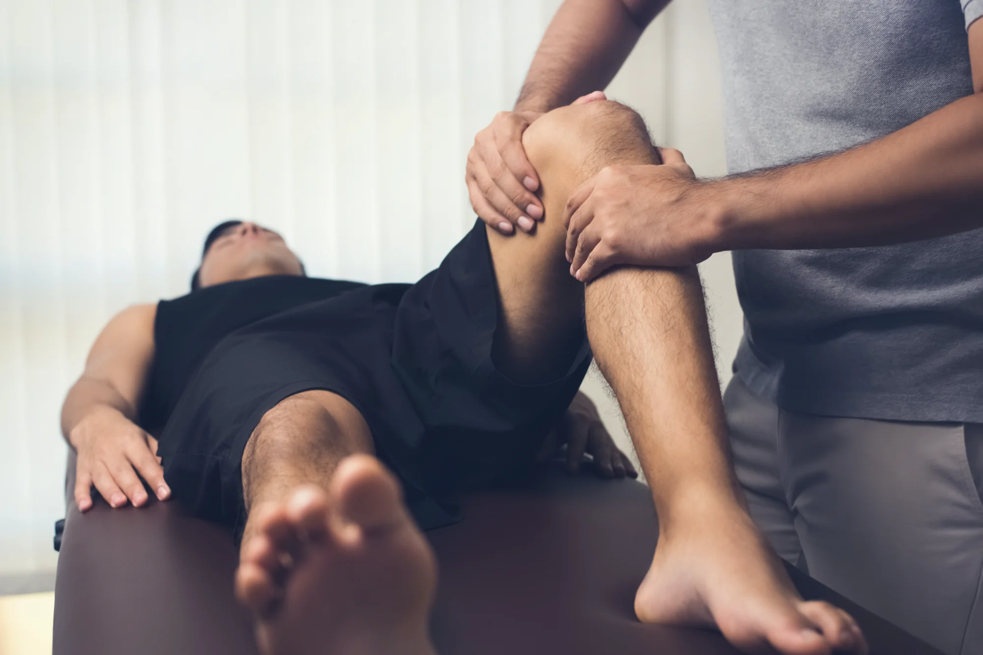 A chiropractor gently manipulates the knee of a male patient who is lying on a dark treatment table, performing a physical adjustment.
