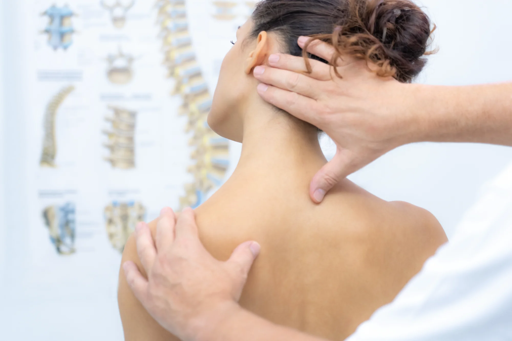A chiropractor adjusts a female patient’s neck and shoulder, with anatomical spine charts visible on the wall behind them.