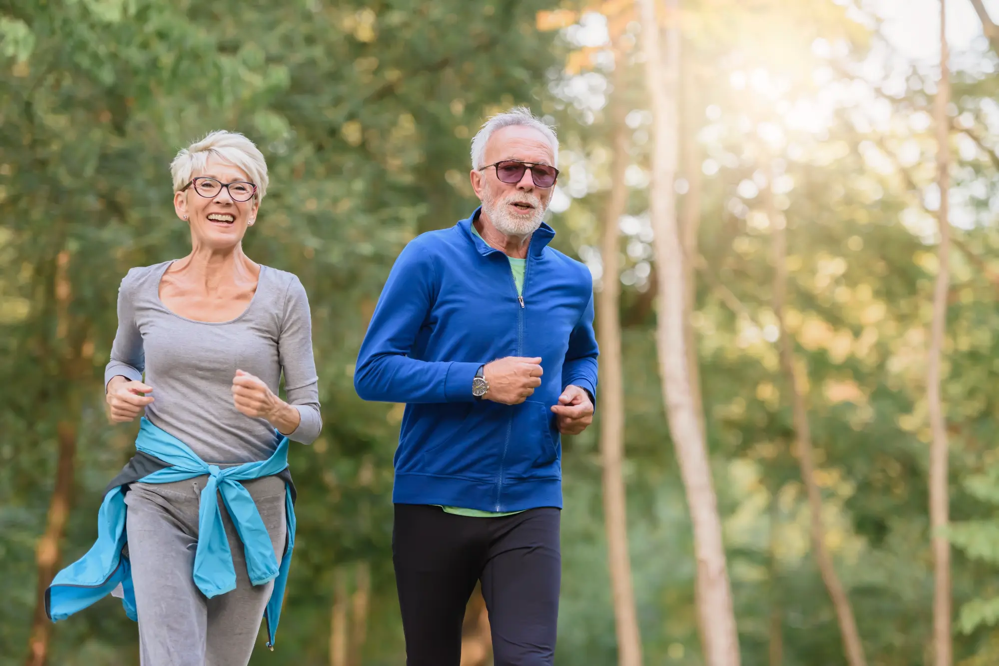 An elderly couple jogging happily outdoors in a sunny wooded area. The woman smiles brightly, while the man with a white beard wears a blue jacket and sunglasses.