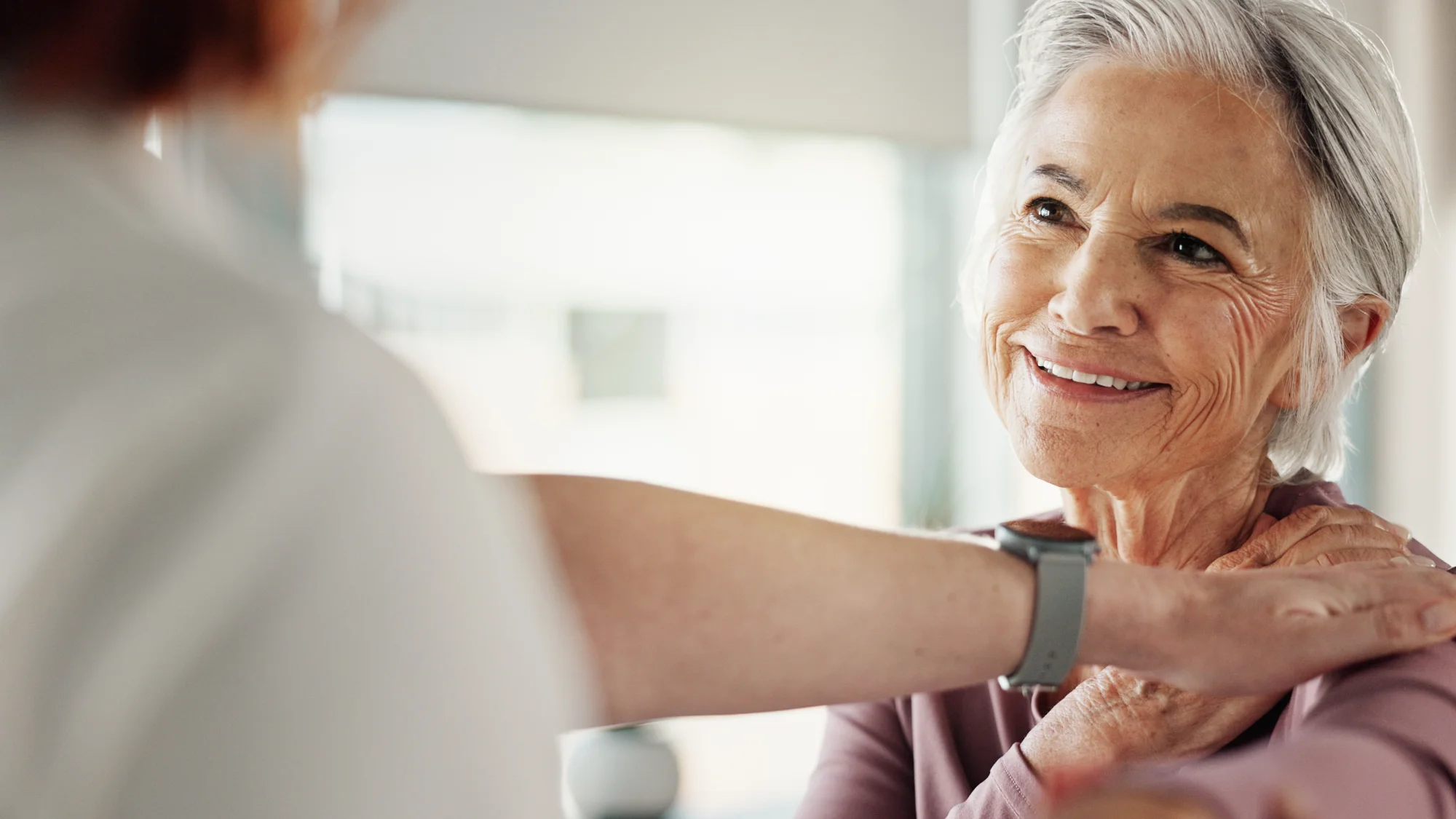 A senior woman with grey hair smiles happily while a medical practitioner, wearing a grey watch, places a supportive hand on her shoulder.