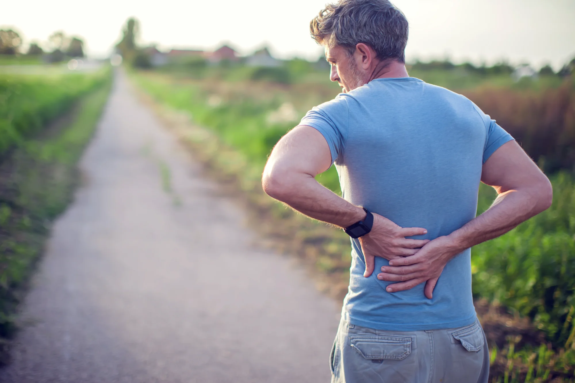 A man in a blue t-shirt stops while walking on an outdoor path, using both hands to grasp his lower back to indicate he is experiencing pain or discomfort.