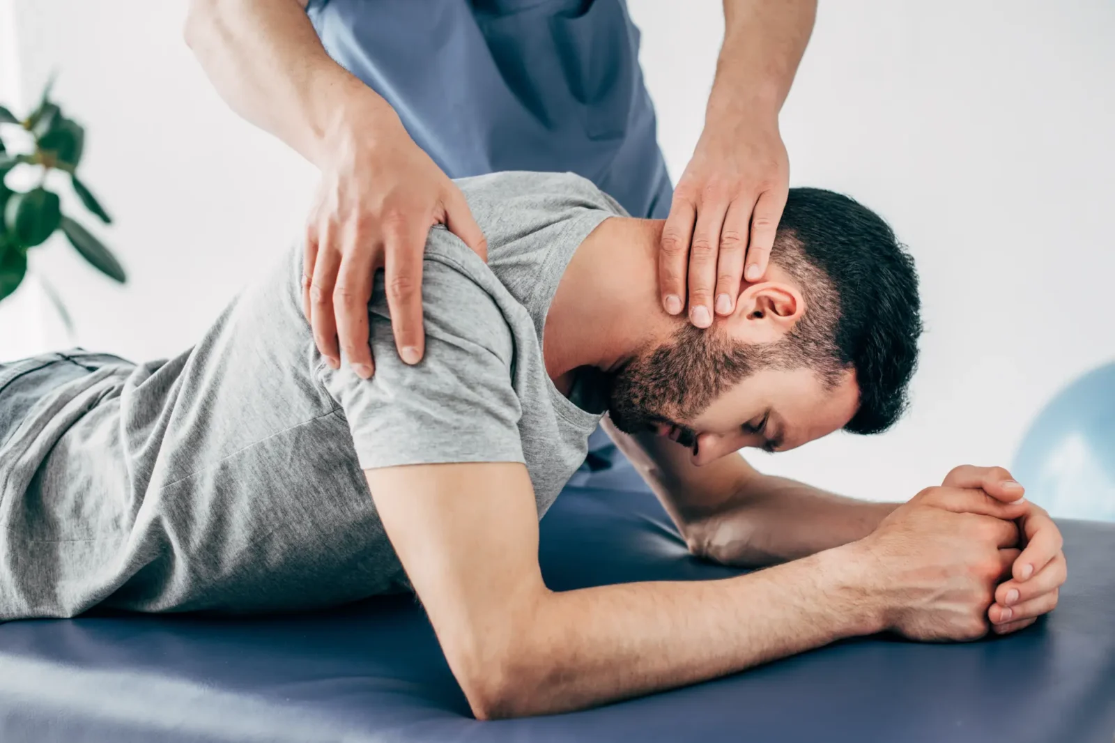 A chiropractor wearing blue scrubs gently adjusts the neck and upper shoulder of a male patient in a gray shirt lying on an examination table.