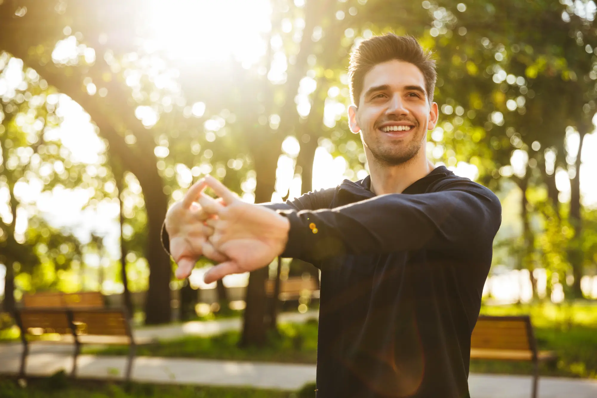 A smiling man in a dark shirt stretches his arms outdoors as bright sunlight shines through the trees behind him.