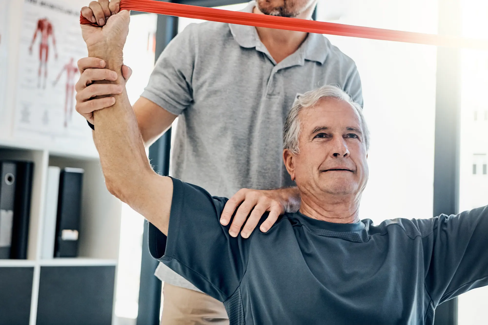 A chiropractor assists an older male patient with an overhead stretch using a red resistance band, supporting his shoulder to guide the movement.