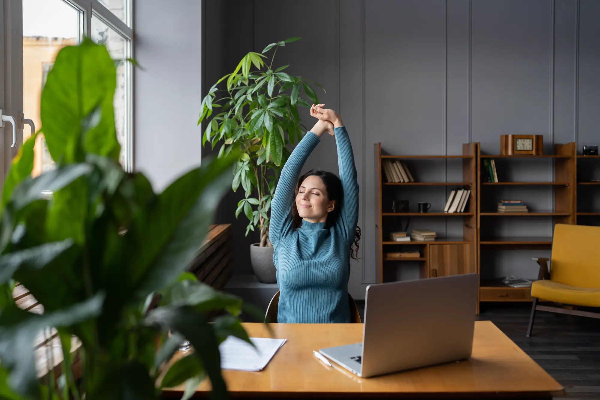 A woman in a blue sweater stretches her arms overhead at her office desk with a relaxed smile and closed eyes, taking a break from her laptop.