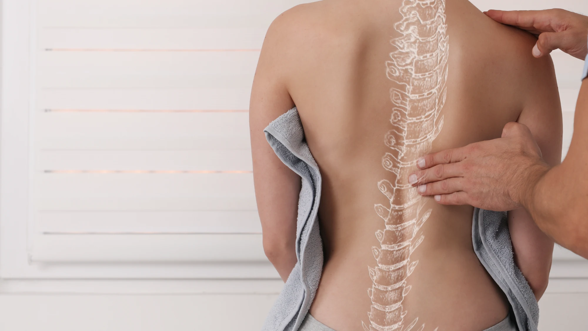 A person's bare back is examined by a practitioner's hands. A white skeletal diagram of the spine is overlaid on the skin for demonstration. The patient is draped in a towel.