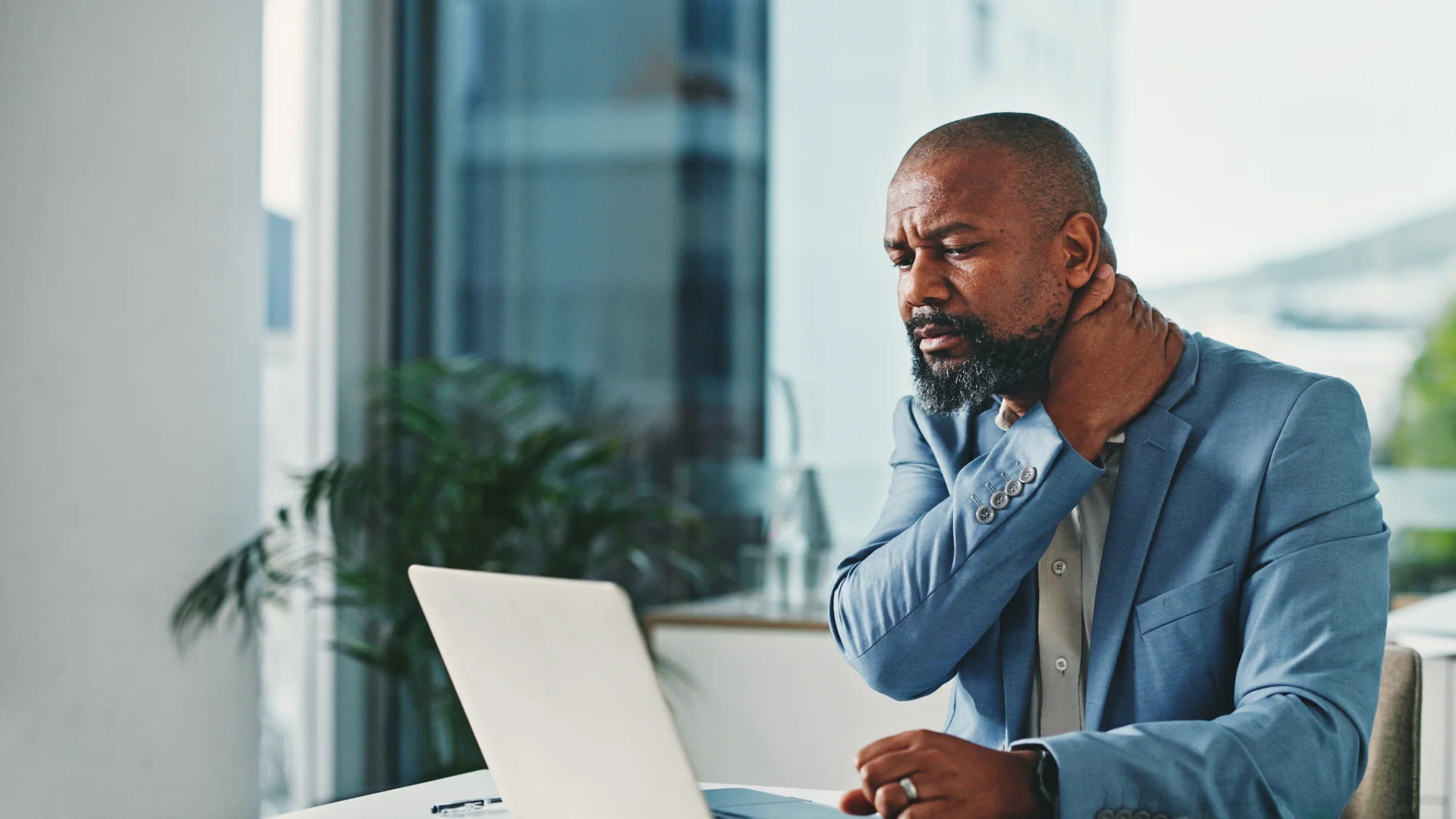 A bearded man in a blue suit sits at his desk with a laptop and rubs the back of his neck with a look of discomfort, suggesting pain or strain.