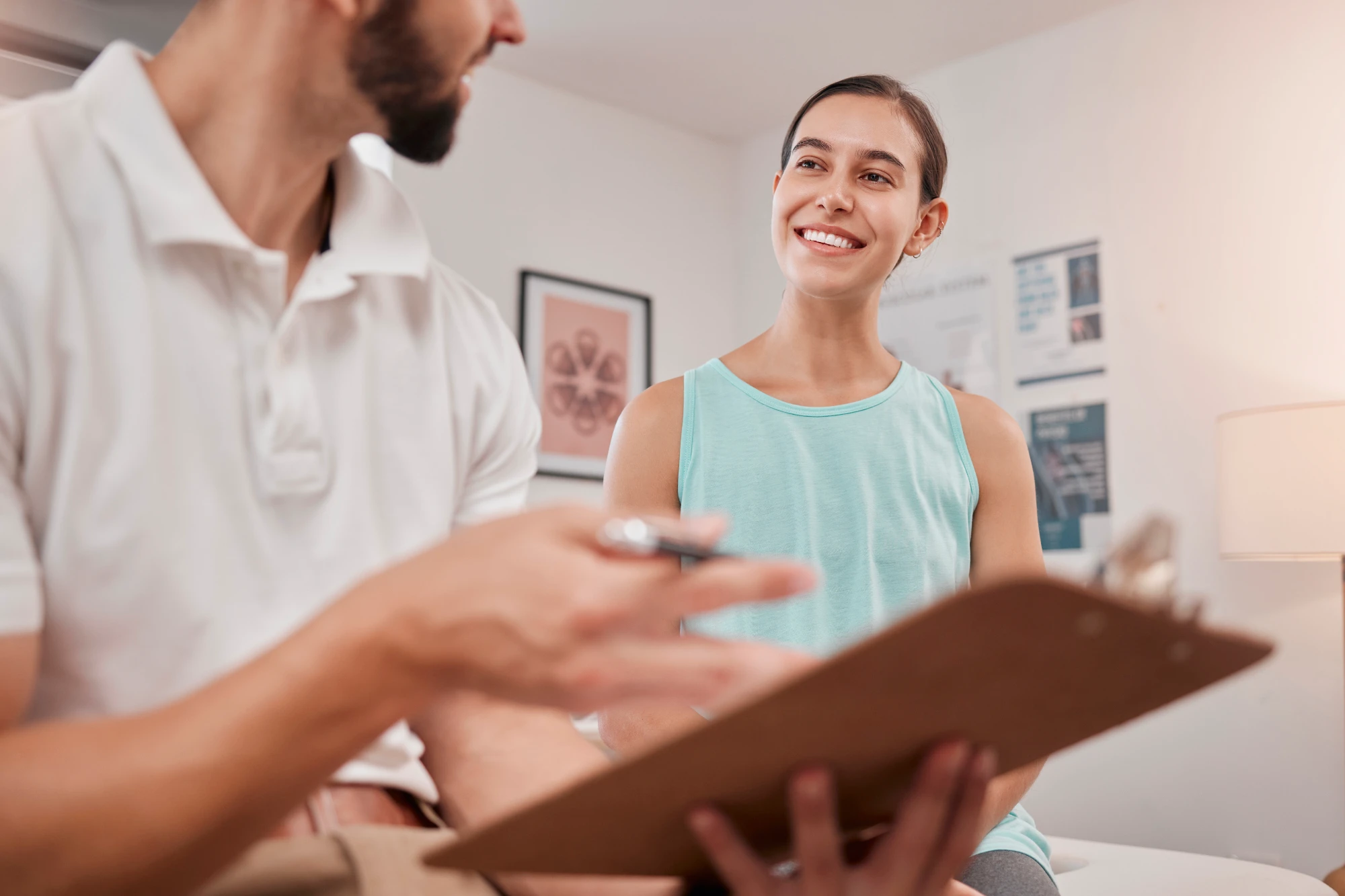 A male chiropractor in a white shirt shows a clipboard to a female patient in a blue tank top who smiles brightly during the consultation.