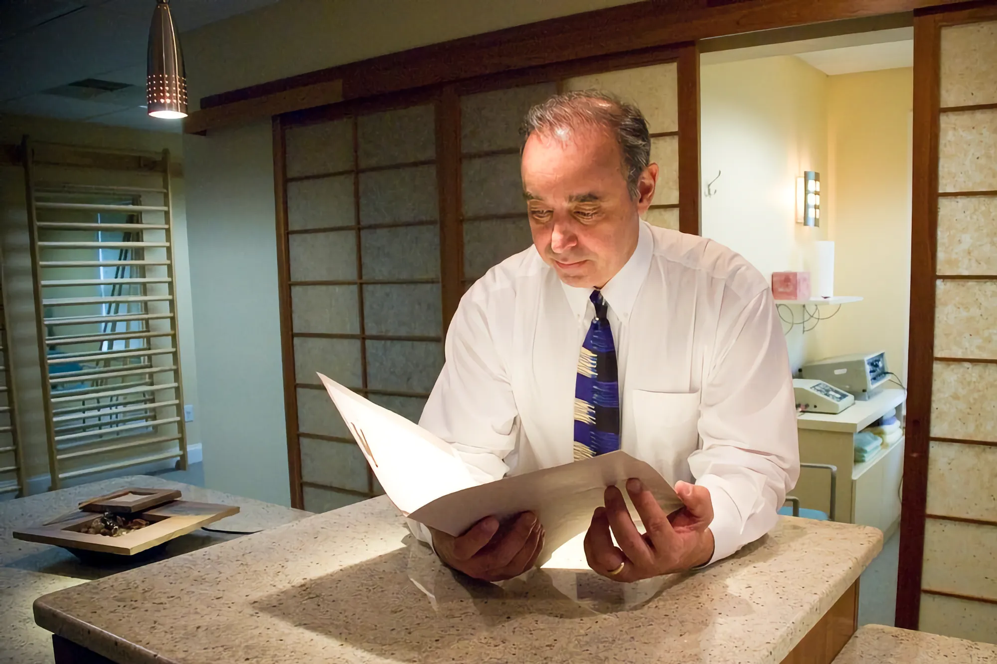 Dr. Marc Moramarco, wearing a white shirt and tie, reviews a patient chart intently at a stone countertop in his softly lit, modern office.