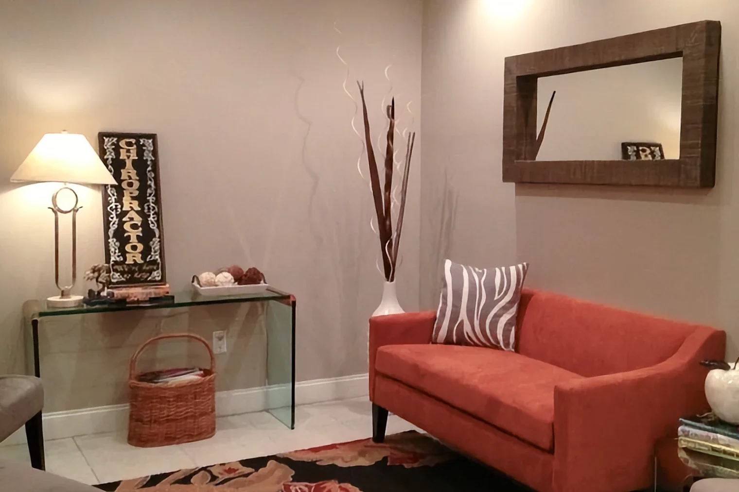 A warm, well-lit chiropractic waiting area corner with an orange loveseat and a glass table displaying a large sign reading "CHIROPRACTOR".