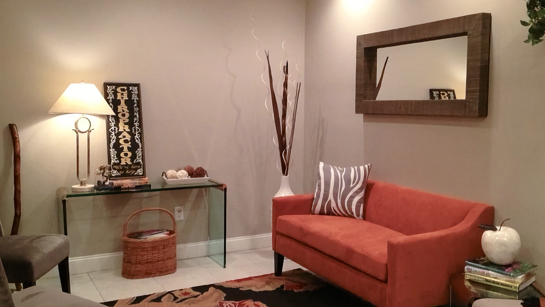 A warm, well-lit chiropractic waiting area corner with an orange loveseat and a glass table displaying a large sign reading "CHIROPRACTOR".