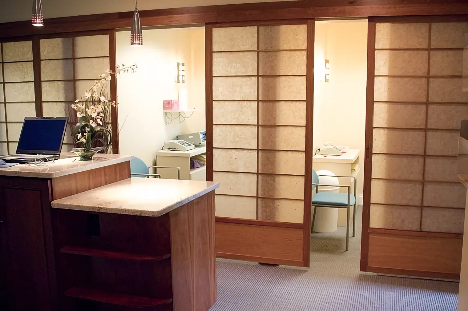 A modern health office uses sliding wooden Shoji screens to partition private rooms containing light blue chairs and medical devices. A laptop rests on the reception counter.