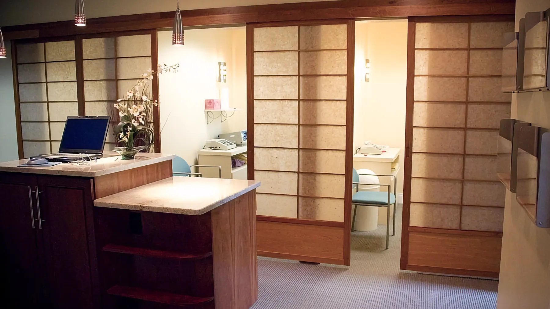 A modern health office uses sliding wooden Shoji screens to partition private rooms containing light blue chairs and medical devices. A laptop rests on the reception counter.
