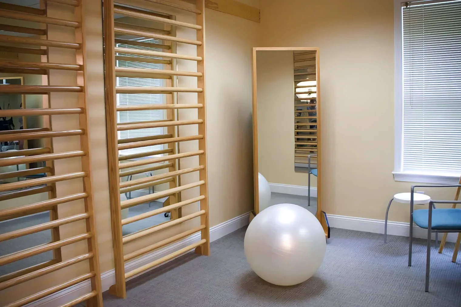 A bright physical therapy room featuring wooden stall bars, a white exercise ball, and a tall mirror on a beige wall.