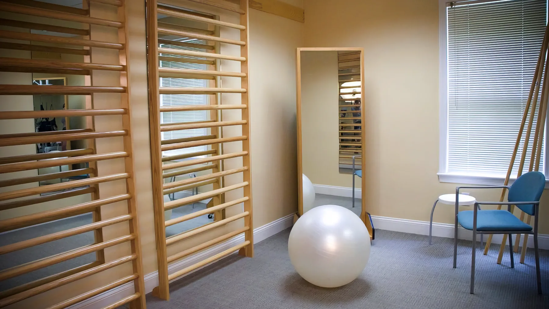 A bright physical therapy room featuring wooden stall bars, a white exercise ball, and a tall mirror on a beige wall.