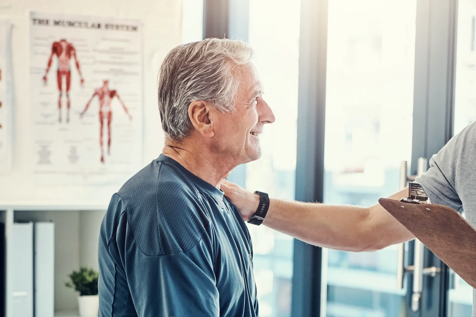 A smiling older male patient in a blue shirt is evaluated by a chiropractor touching his shoulder, with a muscular system chart on the wall.