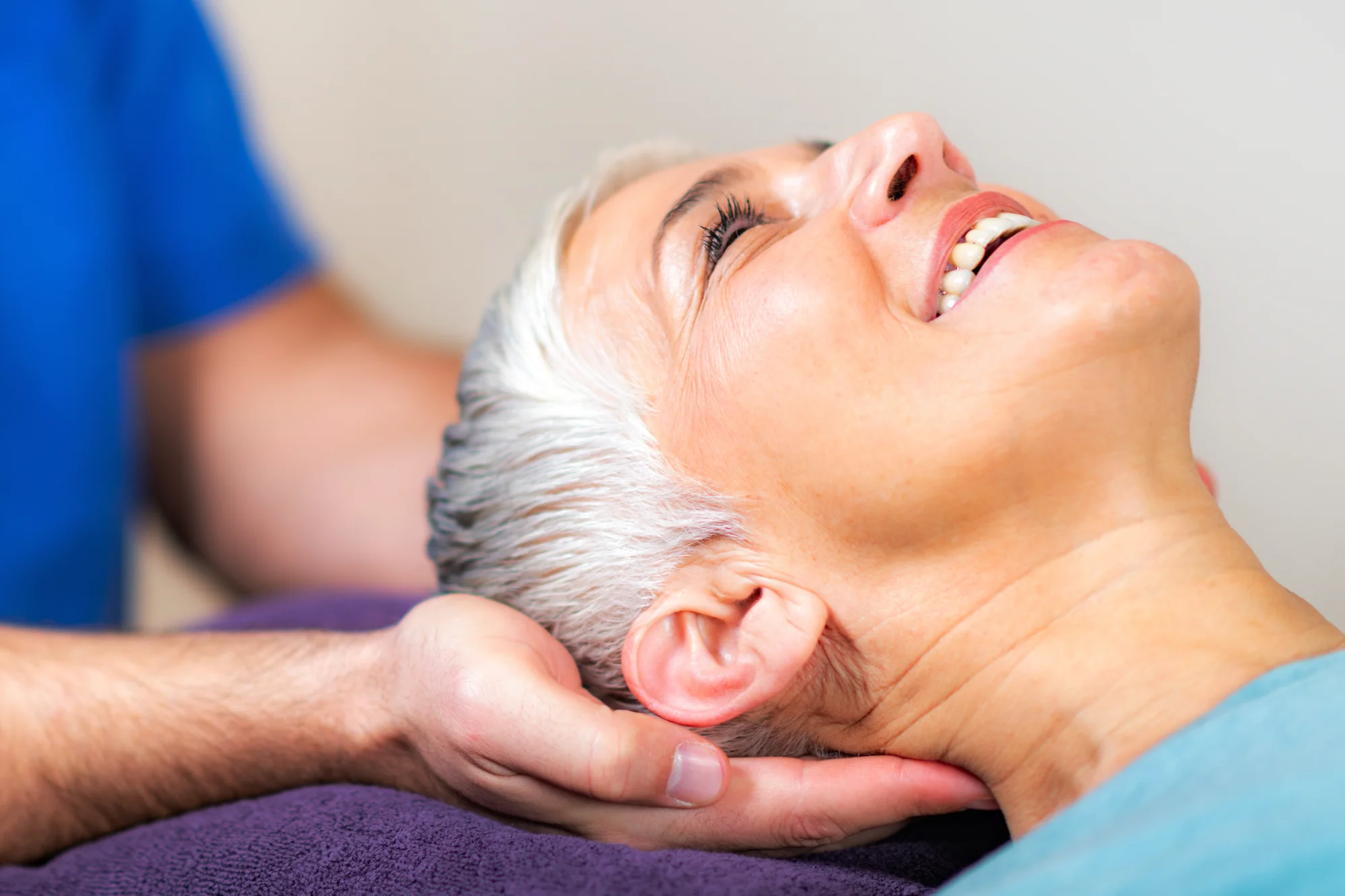 A patient with gray hair smiles widely as a practitioner supports their head and neck with both hands during an evaluation or adjustment.