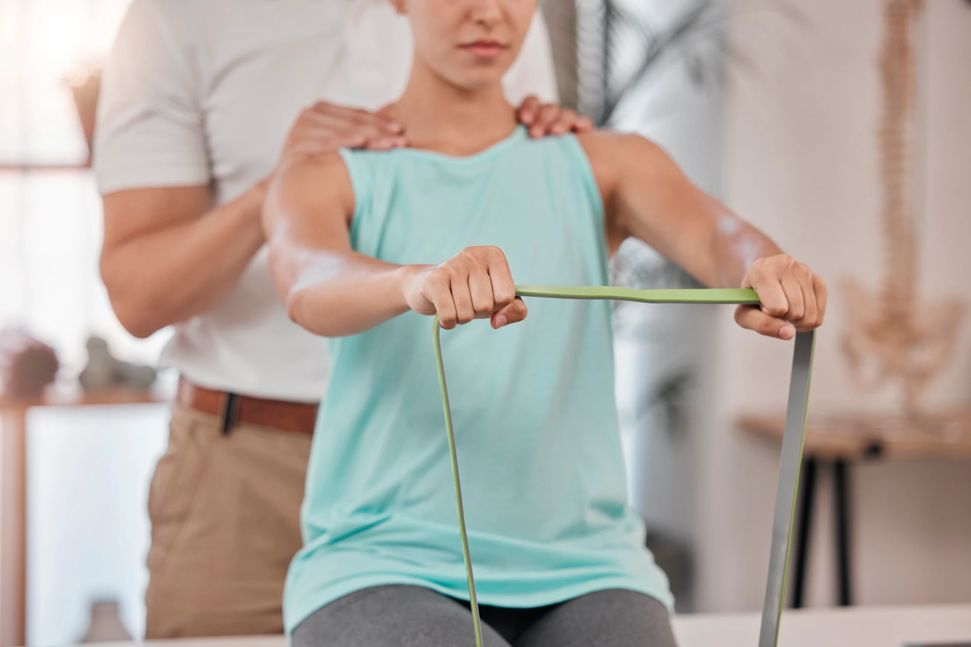 A person in a white shirt places hands on the shoulders of a focused client who is pulling a green resistance band during physical therapy.