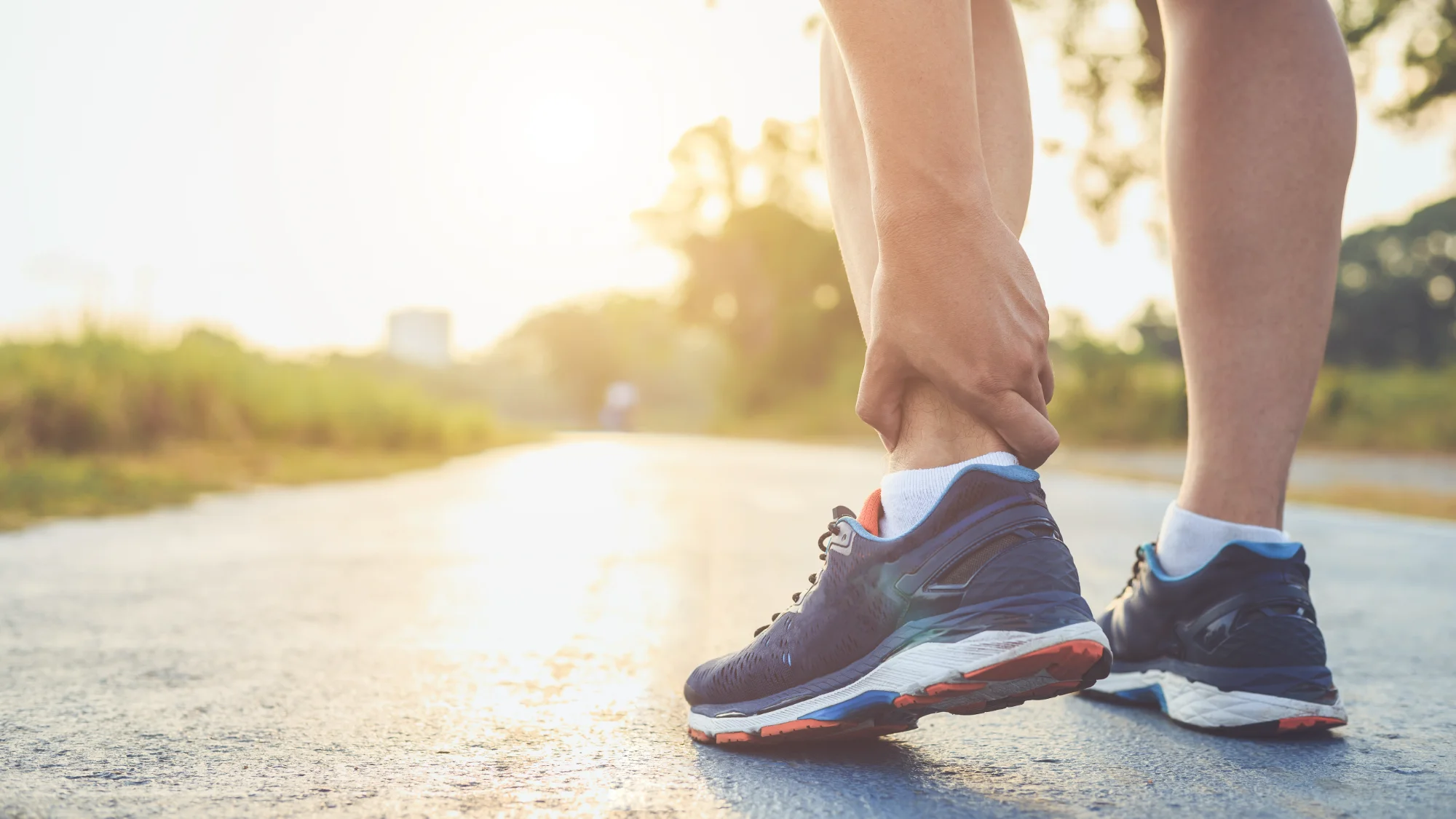 A person in blue running shoes grasps their injured ankle while standing on a sunlit road or trail, suggesting a painful running injury.