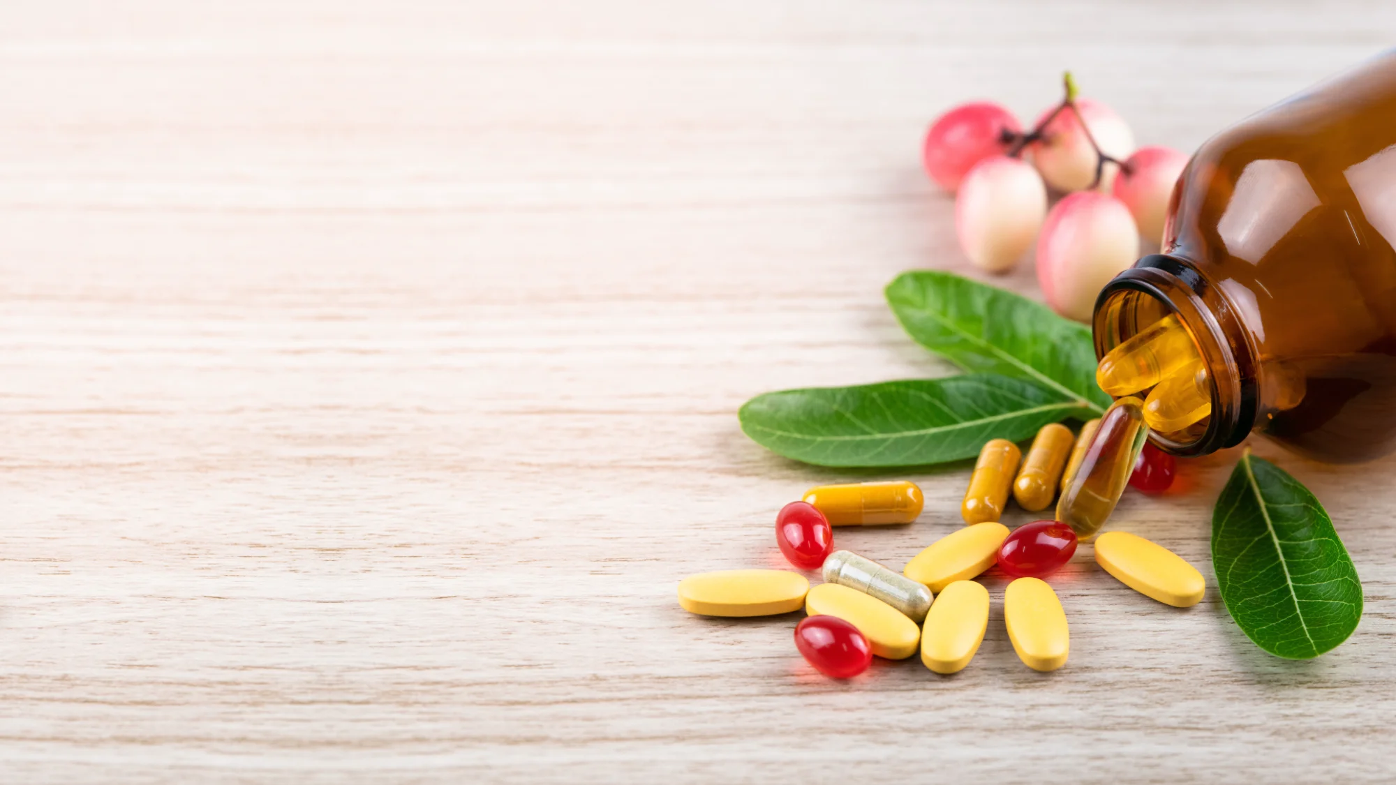 A dark amber bottle is tipped over, spilling various yellow, orange, and red vitamin pills onto a light wooden surface, next to green leaves and some pink berries.
