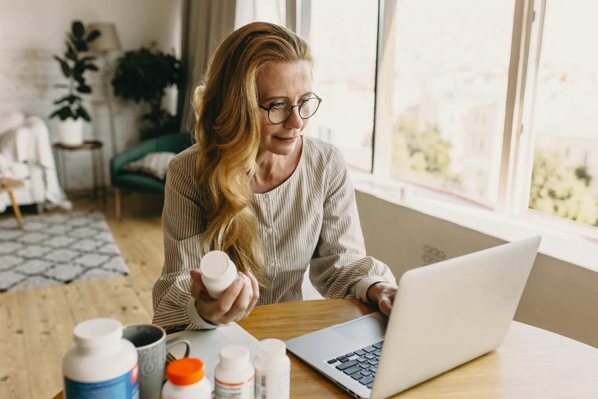 A woman with blonde hair and glasses sits at a desk, looking at her laptop. She holds a white bottle of supplements while researching health information online.