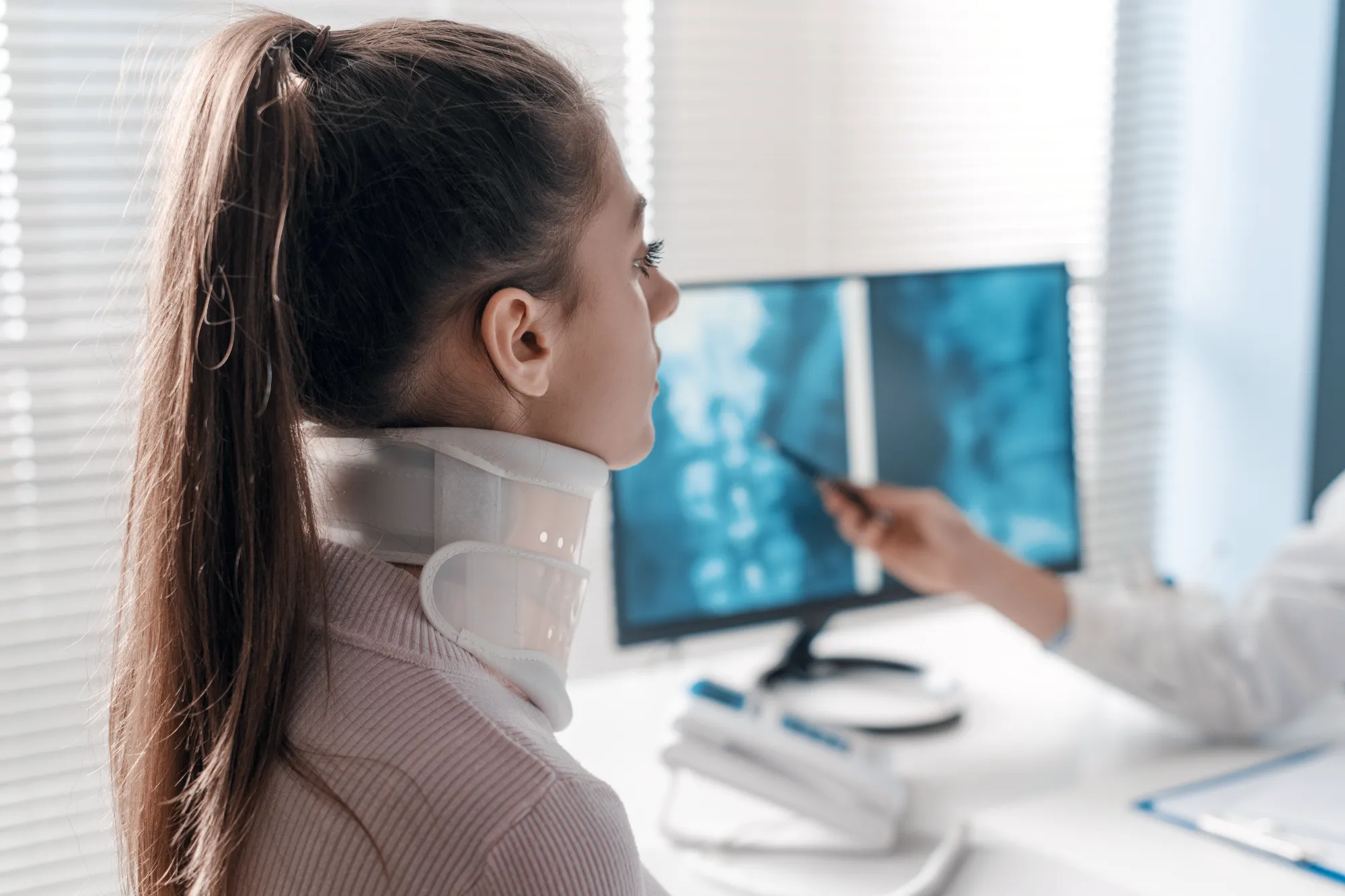 A patient wearing a white cervical collar looks on as a medical professional points to her spine X-rays displayed on a computer screen in an office.