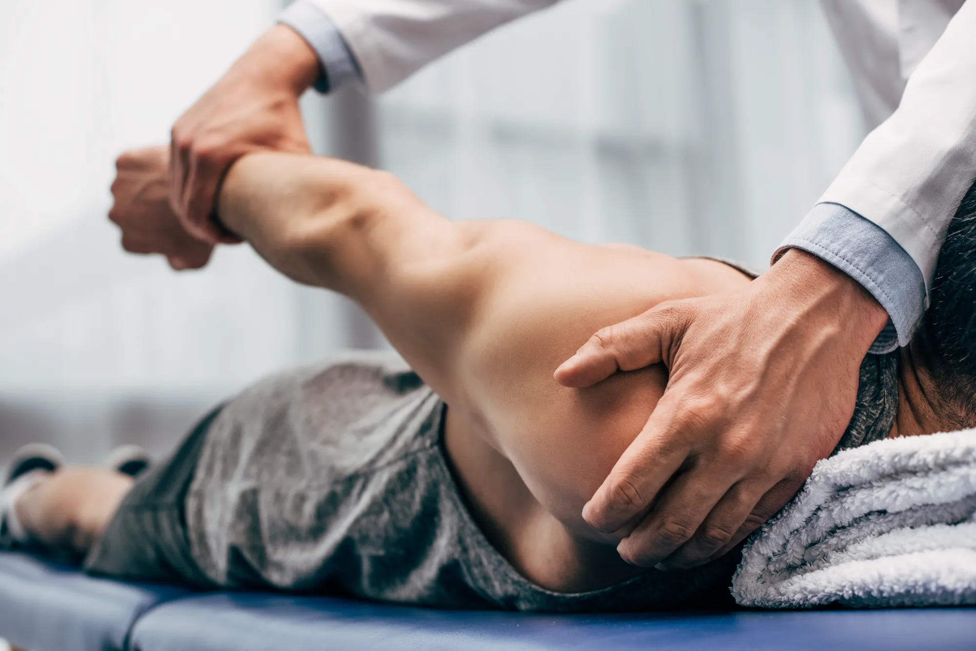 A chiropractor in a white coat performs a shoulder adjustment on a patient lying prone on a blue medical table.