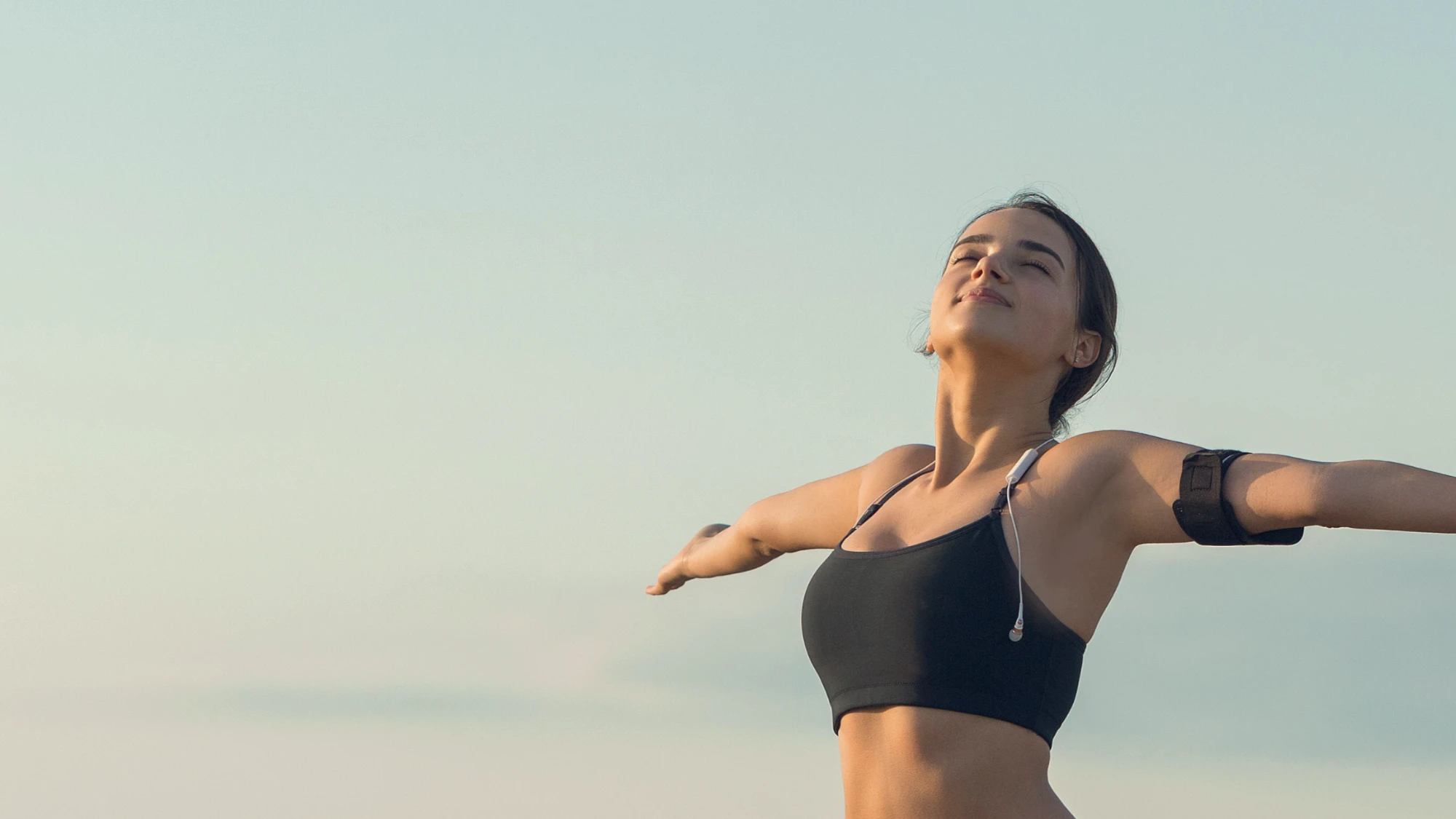 A female runner wearing a black sports bra stands outdoors with her eyes closed and arms outstretched, taking a deep breath against a clear sky.