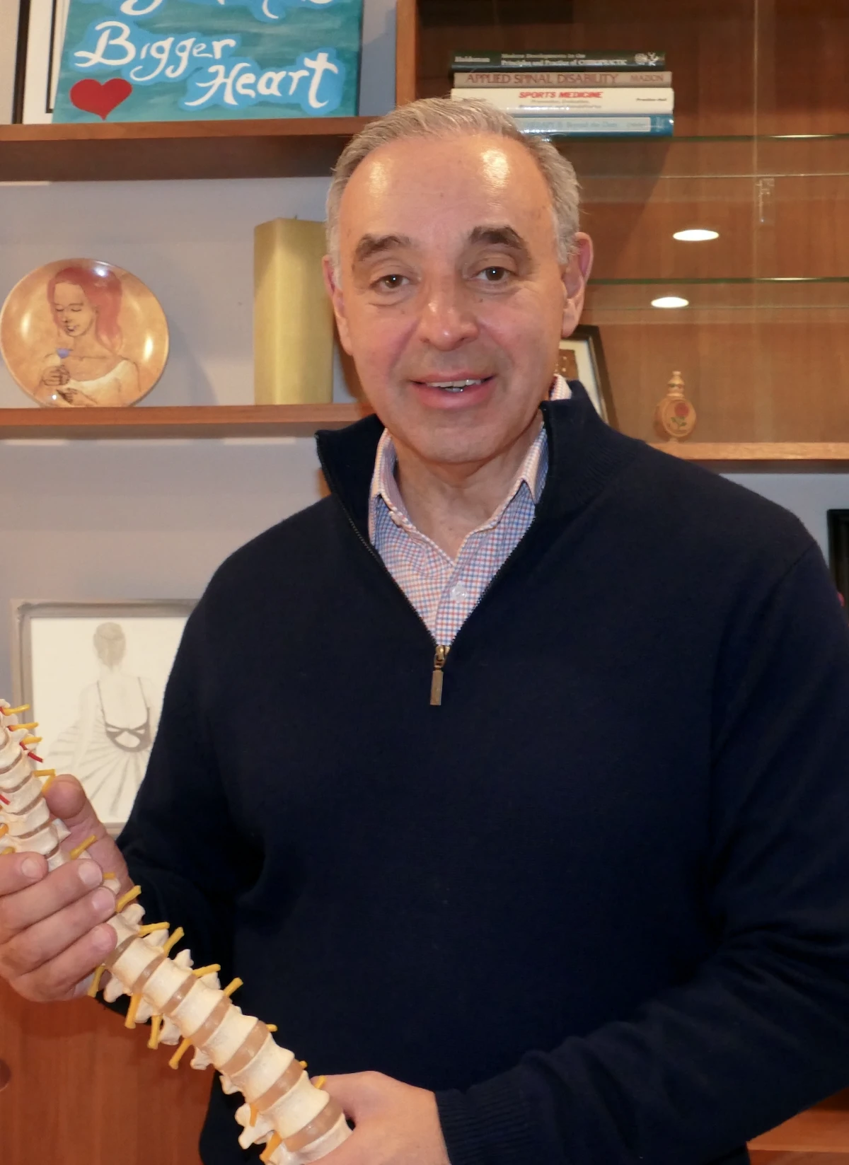 Doctor Marc Moramarco, with grey hair, wearing a navy quarter-zip and plaid shirt, smiles while holding a human spine model in his office, with books on spinal health behind him.
