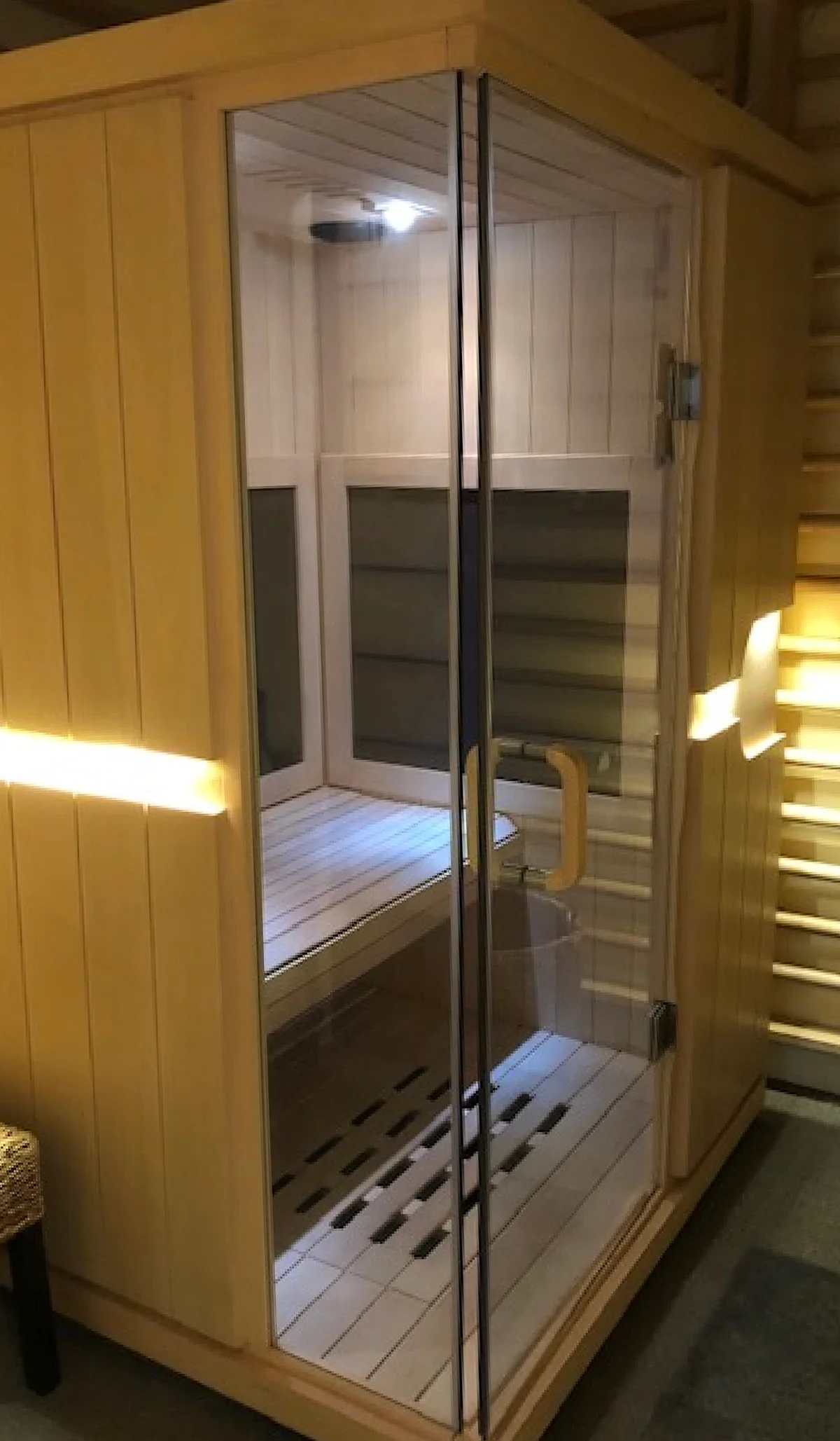 A light wood infrared sauna features a glass door with a wooden handle, an interior bench, a slatted floor, and an internal light, set within an office environment.